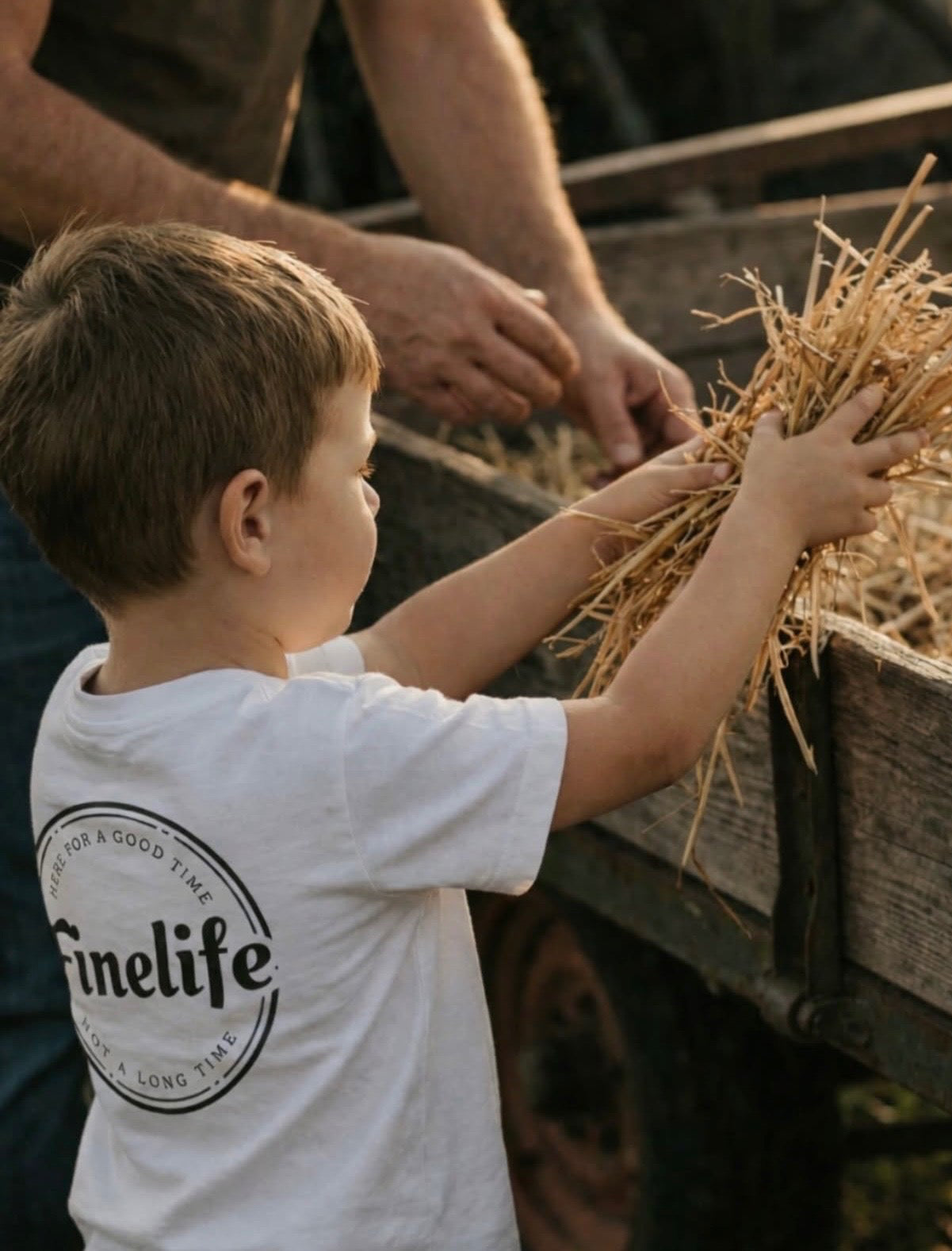 Child in a white 'finelife' shirt holding hay next to an adult on a farm.